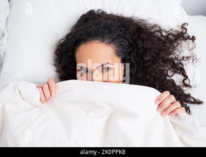 Aufnahme einer jungen, lockigen Frau, die sich unter einer weißen Bettdecke im Bett versteckt Stockfoto