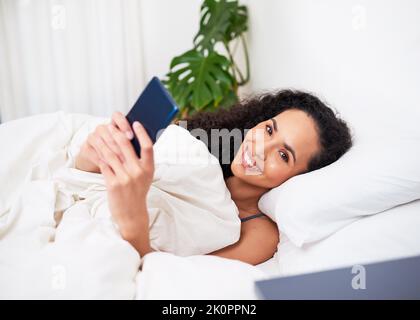 Eine junge, multiethnische Frau liegt lächelnd im Bett und benutzt Handy Stockfoto