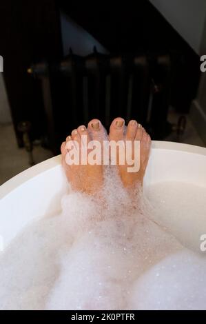 Die reife Frau in ihrem 60s. Alter genießt ein intelligentes, eigenständiges Bad mit ihren Füßen, die mit Seifenblasen bedeckt sind Stockfoto