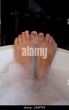 Die reife Frau in ihrem 60s. Alter genießt ein intelligentes, eigenständiges Bad mit ihren Füßen, die mit Seifenblasen bedeckt sind Stockfoto
