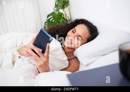 Eine junge, multiethnische Frau liegt lächelnd im Bett und benutzt Handy Stockfoto