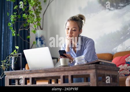 Reife professionelle asiatische Frau sitzt auf der Couch arbeiten von zu Hause aus mit Handy und Notebook-Computer Stockfoto