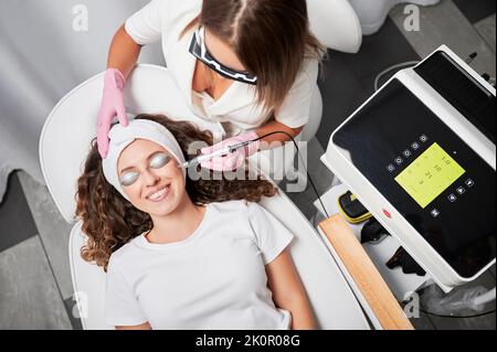 Draufsicht auf glücklichen Patienten mit Augenschutz, der sich im Schönheitssalon einer Laserbehandlung von Gesichtsgefäßen unterziehen muss. Die Kosmetologin entfernt schmerzlos und schnell Teleangiektasen aus dem Gesicht. Stockfoto