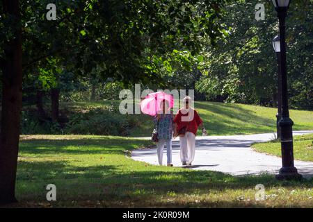 An einem sonnigen Spätsommertag machen zwei asiatische amerikanische Frauen mittleren Alters einen Übungsspaziergang. Man beschirmt sich vor der Sonne mit einem hellmagentafarbenen Schirm. Stockfoto