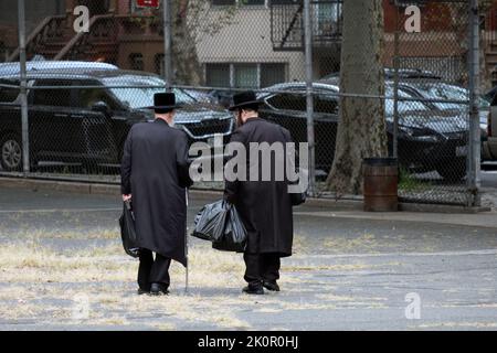 Zwei chassidische jüdische Männer, möglicherweise Familienmitglieder, kehren vom Einkaufen nach Hause zurück und machen eine Abkürzung durch einen Schulhof. In Williamsburg, Brooklyn, New York. Stockfoto