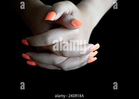 Selektiver Fokus der verflochtenen weiblichen Hände mit orangen falschen Nägeln, auf schwarzem Hintergrund. Nagelmaniküre Stockfoto