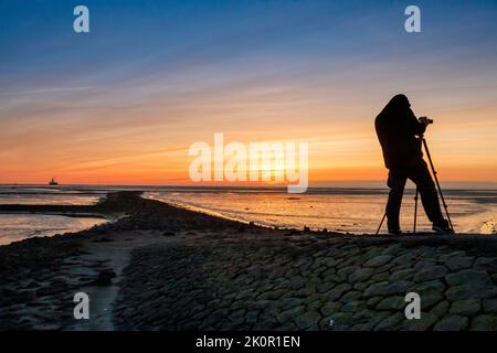 Tourist mit Kamera bei Sonnenuntergang am Tischendamm, Nordsee bei Ebbe, auf der linken Seite die Ölbohrplattform Mittelplate Stockfoto