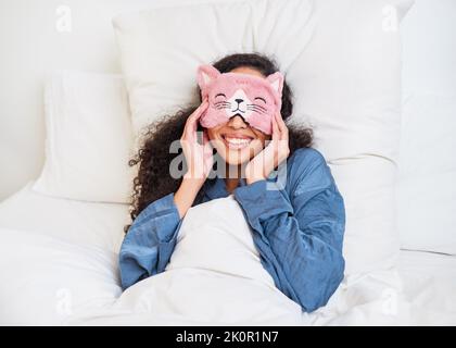 Eine junge multiethnische Frau wacht im Bett mit einer niedlichen Kätzchen-Schlafmaske auf Stockfoto