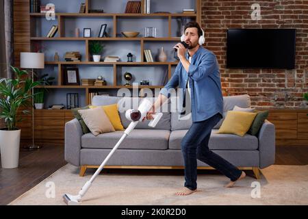 Fröhlicher Kerl, der Mann reinigt die Wohnung. Saugen, Kopfhörer mit Tanz und Gesang in ein Smartphone statt in ein Mikrofon tragen. Haus Stockfoto