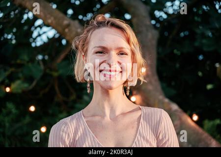 Eine hübsche blonde, reife Frau, die im Garten beim Sonnenuntergang lächelnd auf die Kamera schaut Stockfoto