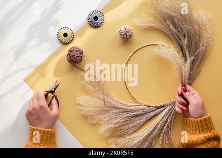 Hände, die getrockneten Blumenkranz aus trockenem Pampagras und Herbstblättern machen. Flach auf weiß gelbem Tisch, Sonnenlicht mit langen Schatten. Stockfoto