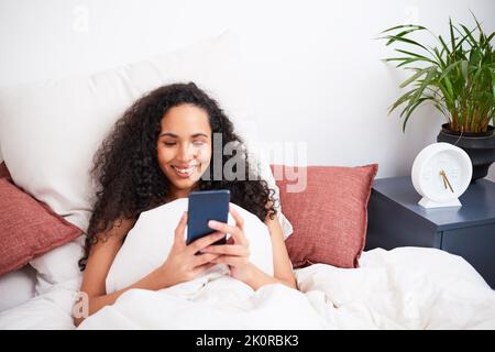 Eine junge, multiethnische Frau sitzt mit dem Handy im Bett mit Kissen Stockfoto