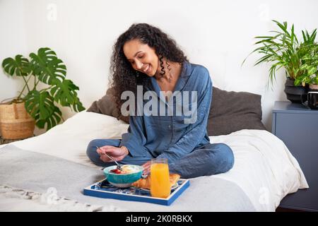 Eine schöne junge Frau isst am Wochenende im Bett in Seidenpyjamas Frühstück Stockfoto