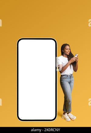 Lächelndes, attraktives Mädchen, das in der Nähe eines riesigen Smartphones mit leerem leeren Bildschirm steht, lächelt, eine Hipster-Frau, die für eine neue App oder Website wirbt, ein Mockup Stockfoto