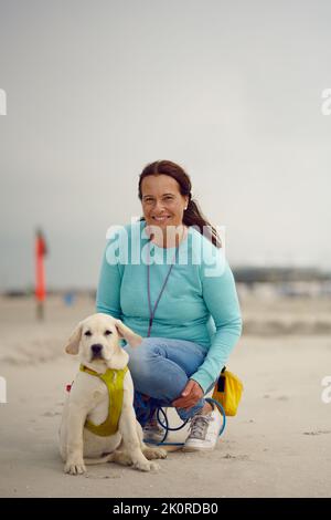 Attraktive lächelnde Frau, die mit ihrem Welpen am Strand posiert und auf dem Sand kniet, mit Kopierfläche darüber Stockfoto