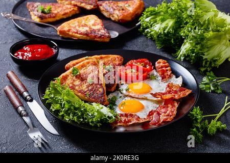 irisches Frühstück mit Spiegeleiern, Speckscheiben, gebratenen Kartoffelkuchen, gegrillten Tomaten und frischem Salat auf schwarzem Teller auf Betontisch mit Zutaten Stockfoto