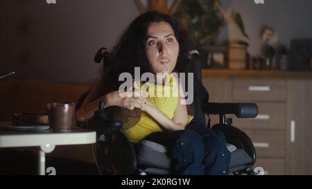 Frau mit spinaler Muskelatrophie sitzt in einem Rollstuhl vor dem Fernseher und schaut sich abends alleine zu Hause einen Film an Stockfoto