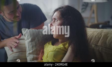 Verwirrte Frau mit einer Behinderung, die auf einer Couch sitzt und mit einem nervösen Mann spricht, während sie streitet und streitet Stockfoto