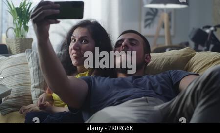 Frau mit einer körperlichen Behinderung und Mann lachen und machen ein Selfie zusammen auf dem Smartphone für soziale Netzwerke sitzen auf einer Couch zu Hause Stockfoto