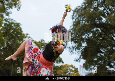 Junge lateinische kaukasische Tänzerin, die im Park mit catrina Make-up lächelt, folkloristische Kleidung trägt, Choreographie übt und Blumen in h hält Stockfoto