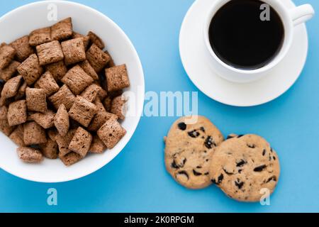 Draufsicht auf Getreideflocken in einer weißen Schüssel in der Nähe einer Tasse Kaffee und Schokoladenkeksen, isoliert auf blau, Stockbild Stockfoto