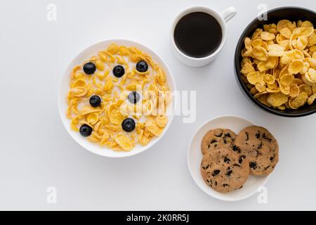 Draufsicht auf Schüsseln mit Maisflocken in Milch mit Heidelbeeren in der Nähe einer Tasse Kaffee und Keksen auf weiß isoliert, Stockbild Stockfoto