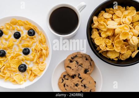 Draufsicht auf Schüsseln mit Maisflocken in Milch mit Heidelbeeren in der Nähe von einer Tasse Kaffee und Schokoladenkeksen auf weiß isoliert, Stockbild Stockfoto