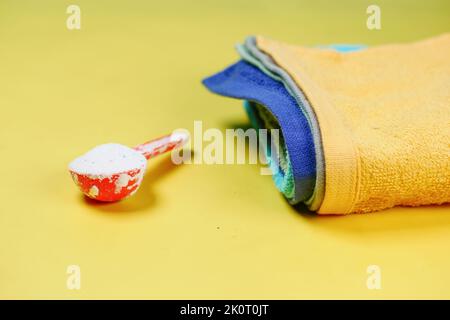 Waschpulver in Plastiklöffel und bunte Tücher auf gelbem Hintergrund Stockfoto