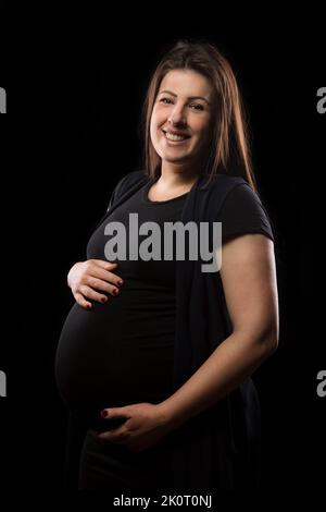 Junge Brünette Frau in späten Stadien der Schwangerschaft lächelt und posiert mit ihrem Baby Beule, Studio Licht verwendet. Stockfoto