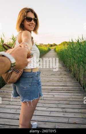 Lässig gekleidete Frau, die bei Sonnenuntergang in der Natur spazierend, meine Hand in der ersten Person haltend und lächelnd. Vertikale Ansicht Stockfoto