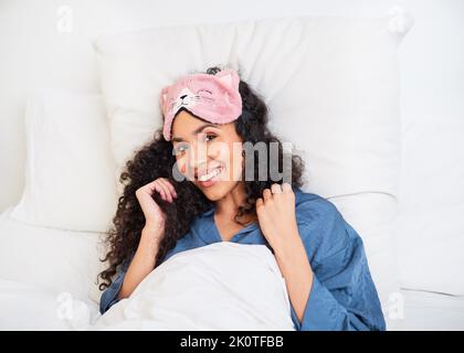 Eine junge multiethnische Frau wacht im Bett mit einer niedlichen Kätzchen-Schlafmaske auf Stockfoto