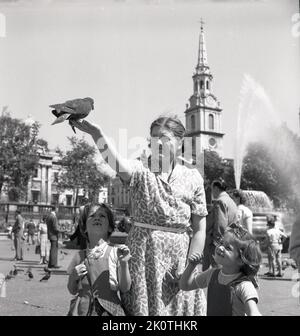 1950er Jahre, historisch, mit ihren beiden jungen Töchtern neben ihr, eine Dame mit einer Taube sitzt auf ihrem ausgestreckten Arm am Trafalgar Square, London, England, UK. Eine traditionelle Aktivität seit über hundert Jahren, Scharen von Tauben steigen auf den Platz, um Essen von denen zu erhalten, die das berühmte Wahrzeichen von Central London besuchen. Stockfoto