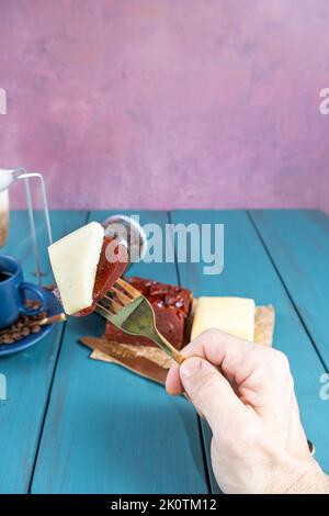 Mann, der eine Gabel mit Guava-Stücken mit Käse hält. Im unscharfen Hintergrund Bohnen und Kaffeetasse vertikal. Stockfoto