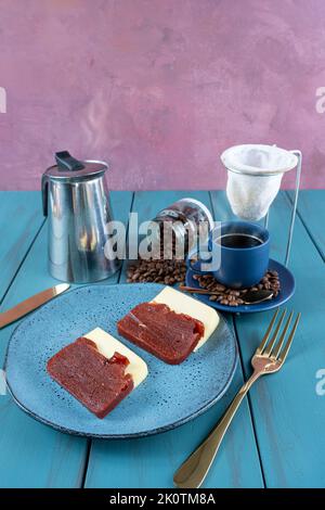 Guava süß mit Käse, umgeben von Besteck, Tasse und Kaffeebohnen auf einem blauen Tisch vertikal. Stockfoto