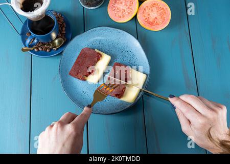 Frau, die Besteck verwendet, um Guava süß mit Käse zu schneiden, umgeben von Bohnen und Kaffeetasse. Stockfoto