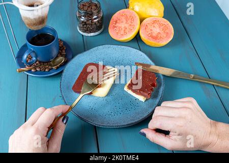 Frau, die eine Gabel über Guava-Scheiben mit Käse hält, umgeben von Bohnen und einer Tasse Kaffee. Stockfoto