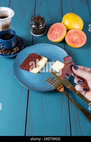 Frau hält eine Scheibe Guava süß mit einem Biss, neben Käse, Bohnen und einer Tasse Kaffee vertikal. Stockfoto