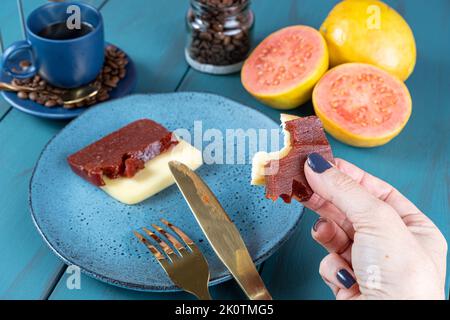 Frau hält Guava-Süssscheiben mit Käse und einem Biss, neben Bohnen und einer Tasse Kaffee. Stockfoto