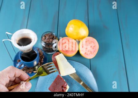 Mann, der eine Gabel mit Guava-Süssstücken mit Käse hält, neben Bohnen und einer Tasse Kaffee. Stockfoto