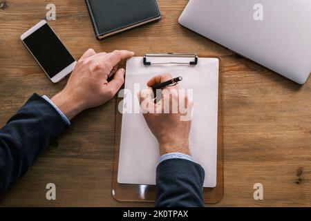 Draufsicht auf männliche Hände, die etwas in einem leeren Briefkopf schreiben, der an eine Zwischenablage geklemmt ist. Geschäftsmann, der am Tisch neben Laptop, Notizblock und arbeitet Stockfoto