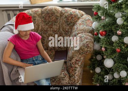 Ein glückliches kleines Mädchen öffnet zu Hause unter dem Weihnachtsbaum ein Geschenk und kommuniziert mit ihren Großeltern über einen Laptop via Videolink. Urlaubskonzept Stockfoto