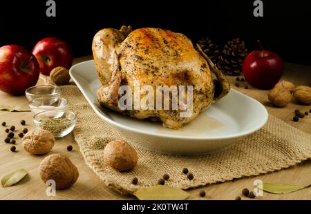 Gebratenes Hähnchen auf weißem Teller, umgeben von Äpfeln, Zapfen, Nüssen und Gewürzen. Saisonales Thanksgiving-Abendessen im Herbst oder Herbst auf Holztisch. Stockfoto