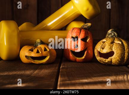 Kleine Halloween-KeramikKürbisse mit zwei gelben Gym-Kurzhanteln. Gesunde Fitness-Lifestyle Herbstzusammensetzung auf Holzhintergrund. Stockfoto