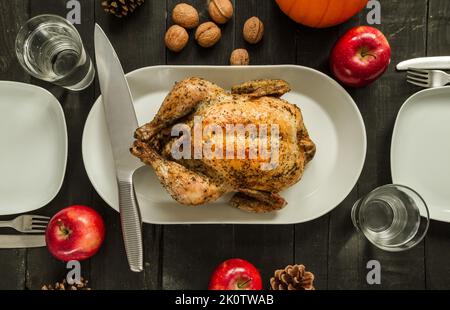 Gebratenes Hähnchen oder truthahn, fertig zum Schnitzen. Thanksgiving- oder Weihnachtsplatten liegen auf Holztisch, mit Äpfeln, Walnüssen, Zapfen und Kürbis. Stockfoto