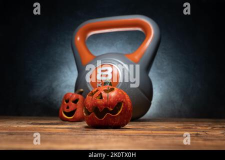 Kleine Halloween-Keramik-Kürbisse mit schwerer Kugelhantel im Hintergrund. Fitness im Herbst oder Herbst mit dekorativen, gruseligen Jack-o'-Laternen. Stockfoto