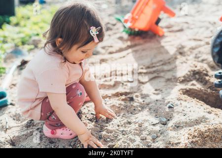 Baby Mädchen sammeln Steine aus dem Sand beim Spielen im Hof des Hauses. Sommer Natur. Schönes junges Mädchen. Glückliche Familie, Kindheit. Stockfoto