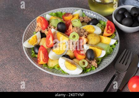 Französischer Salat Nicoise mit Thunfisch, gekochten Kartoffeln, Ei, schwarzen Oliven, Gurken, Tomaten und Salat auf braunem Hintergrund Stockfoto