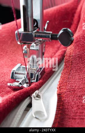 Moderne Nähmaschine mit speziellem Druckfuß näht am Reißverschluss auf rotem Kleidungsstück. Nähprozess Stockfoto