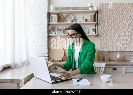 Termin. Eine schöne junge Geschäftsfrau mit Brille sitzt am Tisch in der Küche, arbeitet mit einem kaputten Laptop, eine Computerstörung, winkt ihre Hände unglücklich, zeigt auf den Monitor. Stockfoto