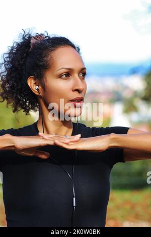 Nahaufnahme Frau im Freien mit Ohrhörern und lockigen Haaren Hände auf Brust Sport-Outfit Stockfoto